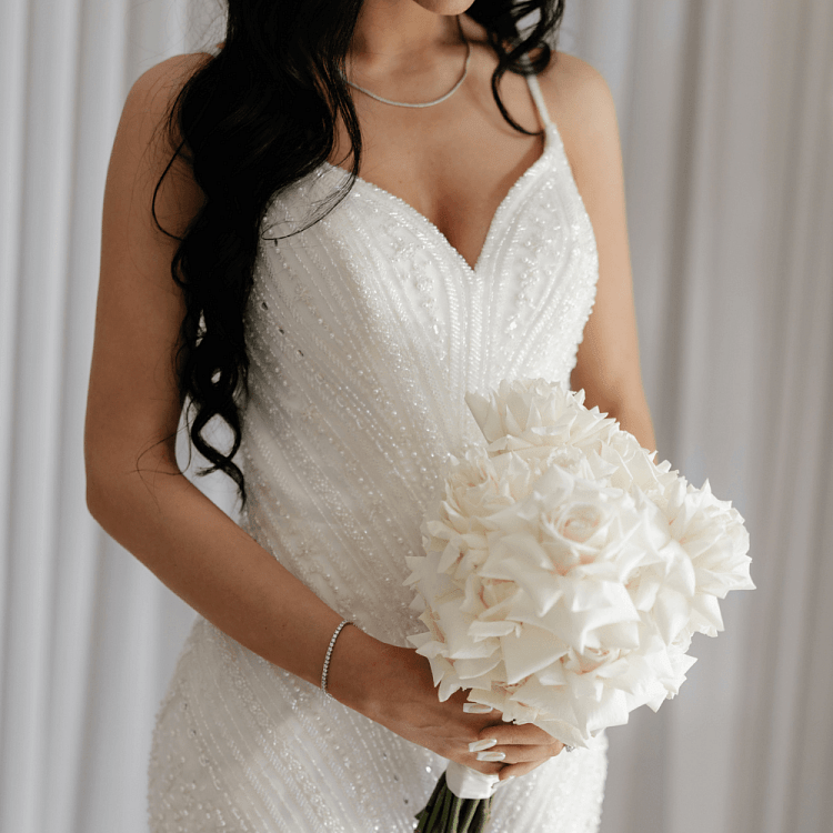 Bride in a beaded white gown holding a bouquet, wearing a simple diamond necklace and bracelet in an elegant close-up.