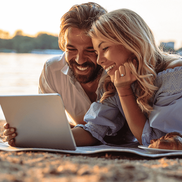 Smiling couple lying on the beach looking at a tablet, choosing together the engagement ring she wants
