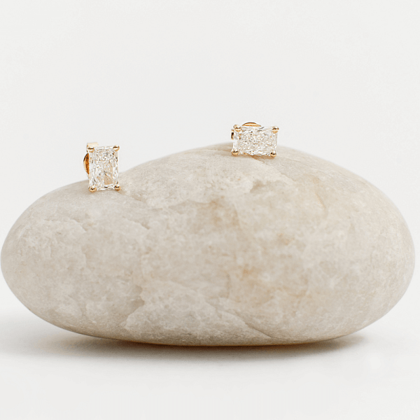 Pair of rectangular diamond stud earrings in gold settings displayed on a smooth white stone.