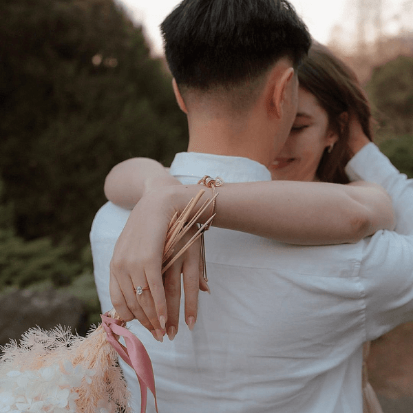Photo of a woman embracing her partner outdoors, showing her engagement ring and holding a bouquet with pink ribbons.