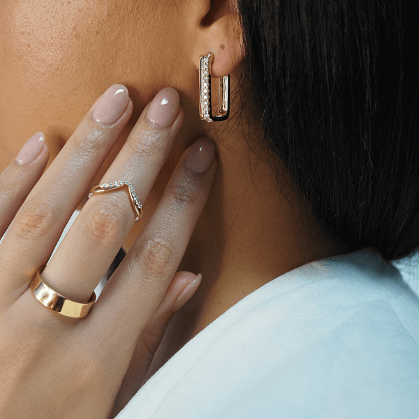 Woman wearing gold hoop earrings and gold rings, including a V-shaped diamond ring and a wide gold band.